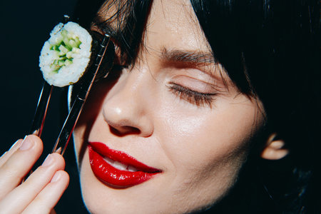 Young woman with black hair enjoying sushi, holding a piece with chopsticks, featuring a close-up expression of delight and vibrant red lips.の写真素材