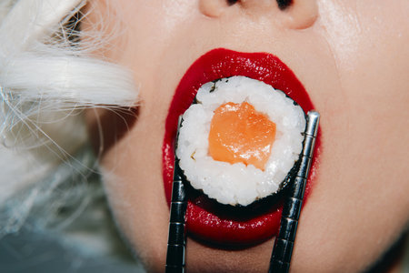Close-up of a womans mouth with sushi and chopsticks, featuring red lipstick and a vibrant sushi roll, capturing the essence of culinary artistry.の写真素材