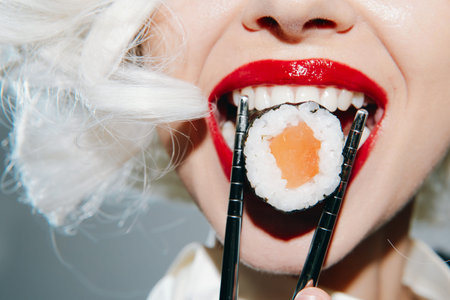 Young woman with red lipstick playfully holding sushi with chopsticks, showcasing a vibrant and...