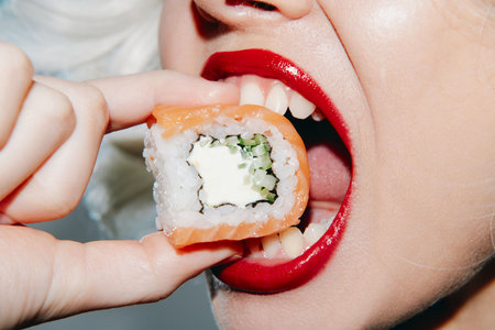 Close-up of a woman's mouth holding sushi, showcasing bright red lipstick and a vibrant sushi roll concept.の写真素材