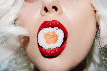 Close-up of a models mouth with sushi, featuring bright red lips and playful expression, emphasizing food aesthetics and vibrant colors.の写真素材