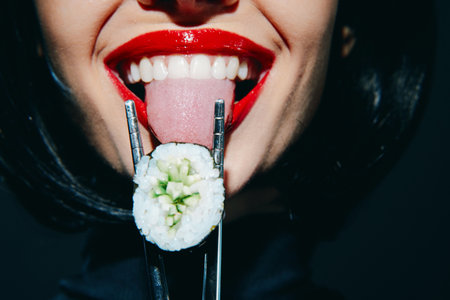 Woman with red lips holding sushi with chopsticks, showcasing emotions against a dark background.の写真素材
