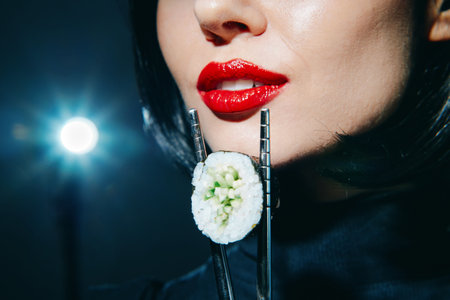 Elegant woman with red lips holding sushi on chopsticks against a dark background, showcasing a glamorous food concept and modern lifestyle.の写真素材