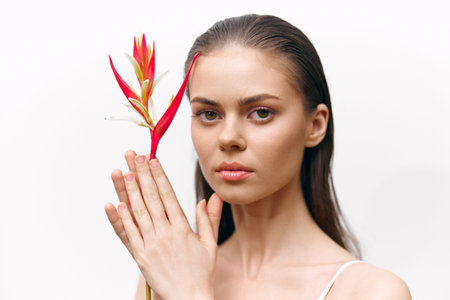 Young woman with long hair holding a vibrant tropical flower against a minimalist white background, conveying serenity and beauty in contemporary fashion.の写真素材