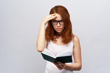 Beautiful redhead young woman with glasses and white shirt on an isolated white background keeps reading a book, she frowns and focuses new information. The student is hard preparing for the exam.の写真素材