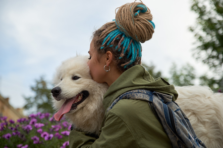 Modern stylish young girl with dreadlocks on her head hugs and kisses her beloved dog snow-white Samoyed. Portrait against the background of spring Park, green flowersの写真素材