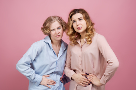 Two young women hold onto their bellies with suffering expression. Studio portrait on pink background. The concept of stomach trouble, menstrual pains, indigestion.の写真素材