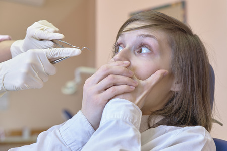 a young white woman in horror looks at the dentist's hands and tools in her hands. In fear, she clamps her mouth with two hands.の写真素材