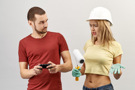 Attractive young woman in jeans, yellow shirt and a hard hat throws up his hands with a disgruntled expression because her male partner is repair and painting playing video games with a joystickの写真素材