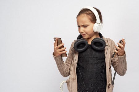 Serious angry girl dressed in warm jacket, presses her lips together and stares fiercely at phone, frowning with displeasure, she wants peace and quiet, two of headphones, three phones and wire.の写真素材