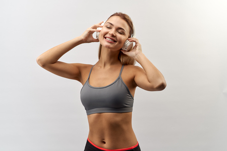 joyful smiling girl in grey sport top with headphones on her head poses for a Studio portrait on a white background with a wide smile and holding hands headphonesの写真素材