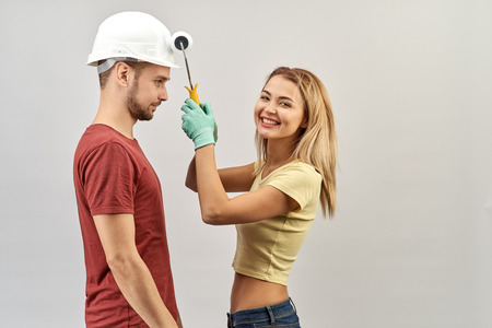 young positive couple fooling around during the repair and painting of the walls in the apartment. A man in a white hard hat and the girl in gloves with paint roller in handの写真素材