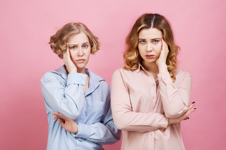 Two young women are upset and holding onto their heads. Headache and anxiety spoil their mood, they feel stress, pain and discontent. Solid color shirt and pink isolated background. Studio portraitの写真素材