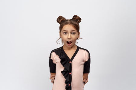 Funny young girl opens wide her mouth. The child is shocked and happy positive news about the birth of a brother or sister in the family. Standing on an isolated white background, pink black sweater.の写真素材