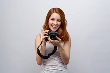 portrait of young attractive redhead girl photographer in a white t-shirt with camera in hand. Joyful expression, wide smile and winks at the camera. Close-up portrait on white backgroundの写真素材