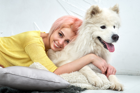 Charming pretty girl looking at camera and hugs her dog, young woman smiles widely and shows beautiful white teeth. A huge white dog looks into distance, his tongue, it hot from the arms of the owner.の写真素材