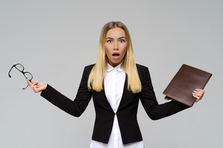 Studio portrait of a young long-haired blonde with an expressive look of surprise opens her mouth and spread her hands to the sides, holding documents and glasses.の写真素材