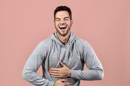 Studio large portrait of a young man laughing happily and holding his stomach from laughter. Snow-white smile and gray hoodie on isolated pink backgroundの写真素材