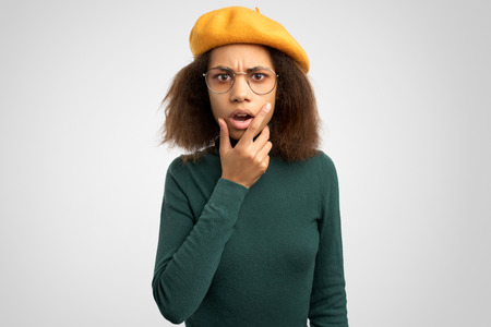 Photo of surprised woman keeps hand on cheek, has dissatisfied expression, recieves bad news, wears sweaters and beret isolated against white background with free space for your text.の写真素材
