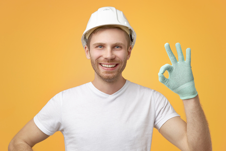 Pleased confident European man with broad smile, uniform, shows okay gesture, dressed in t-shirt and construction helmet. Isolated over white background.の写真素材