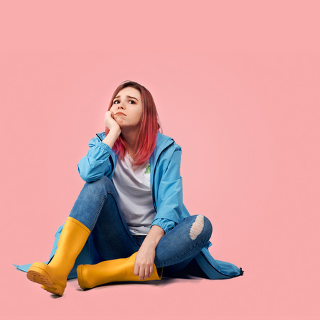 Young beautiful girl in blue coat and yellow rubber boots sits with sad expression in her eyes. She is depressed by spoiled rainy weather. Square shot of woman isolated on pink background in Studio.の写真素材