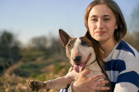 A beautiful woman wearing a striped jumper hugging a bull Terrier dog on a field background. A happy girl with a snow-white smile laughs and enjoys a walk in the open air.の写真素材