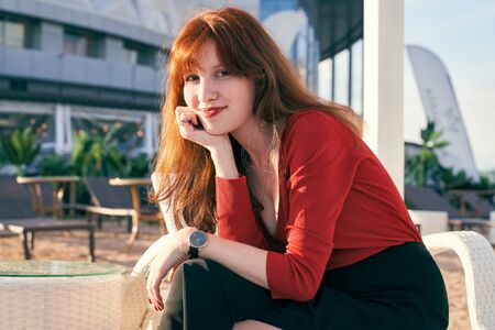 An attractive young girl with long curly redhead hair has bright red lipstick and freckles, wearing jumper and skirt, sitting on white chaise longue on terrace cafe by river. Sunny evening in summer.の写真素材
