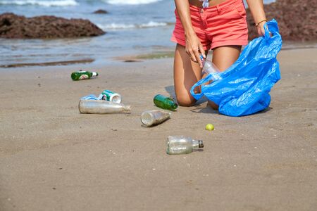 Care about nature. Young woman tourist volunteer busy collecting rubbish, picks up plastic bottles, protects environment from disastrous consequences, holds garbage bag, cleaning beach from trash.の写真素材