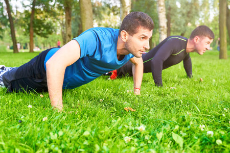 The men team does push-UPS on street on sports ground. Instructor with big biceps teaches lesson on...