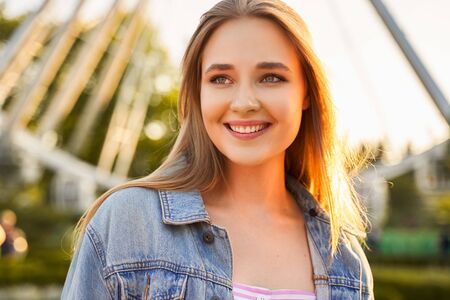 Outdoor portrait of charming European girl, illuminated by pleasant evening sun, looking aside with beautiful joyful smile, dreaming, being inspired and satisfied with summer holiday in city park.の写真素材