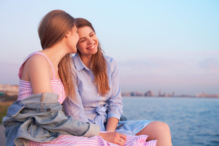 Two excited pretty young girls have warm friendly relationship, sit on sea shore, embrace each other...