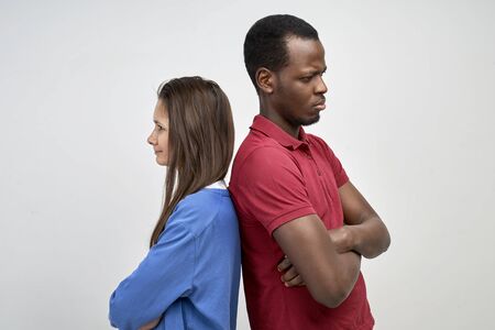 A young African and European couple stand with their backs against the white wall. Two people got into a fight and don't compromise.の写真素材