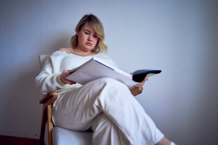 medium-sized woman in light clothes reads a book while sitting in a white chair in a light roomの写真素材