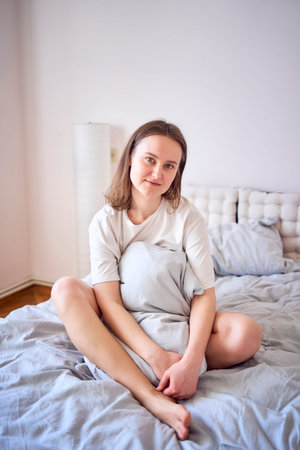 a beautiful young woman in white t-shirt on bed hugging pillowの写真素材