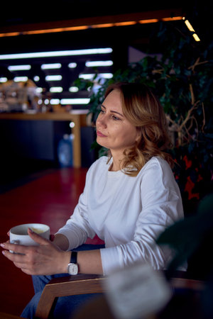 a beautiful middle age woman drinking coffee in a beautiful cafe with greenery and light panels in...