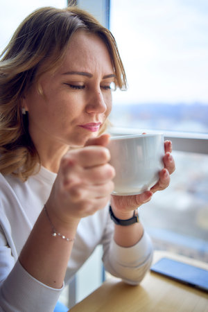 a young blonde woman drinking coffee and working on a laptop in a cafe with panoramic windows and a view of the city from aboveの写真素材