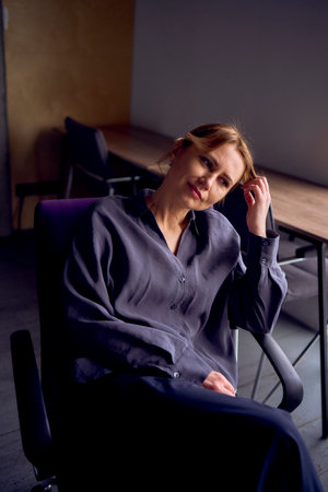 beautiful middle age business woman wearing gray shirt, wide leg pants abd black stilettos sit in purple office chair in modern workspaceの写真素材