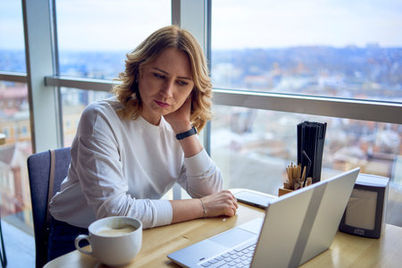 a young blonde woman drinking coffee and working on a laptop in a cafe with panoramic windows and a view of the city from aboveの写真素材