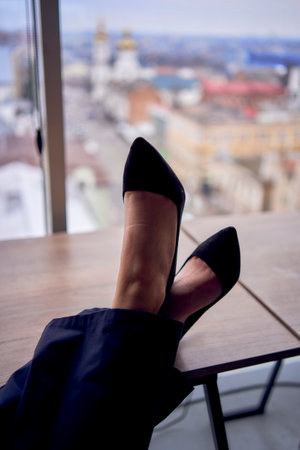 Person sitting in front of window with high heels, black pants and leg crossed. Cityscape outside. Modern interior with neutral tones.の写真素材