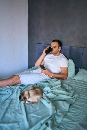 young man drinking morning coffee in bed and scrolling in smartphone, morning routine