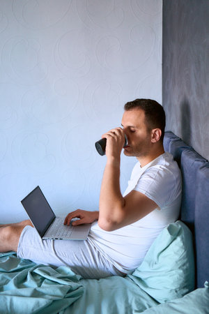 young man drinking morning coffee in bed working on laptopの写真素材