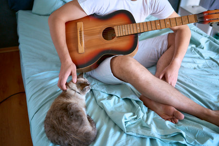 young man is learning to play the guitar at home, a cat is lying next to him and listeningの写真素材