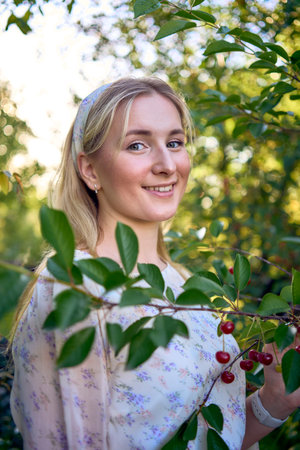 a portrait of a young blonde woman in a pastel chiffon dress among the branches of a ripe cherry