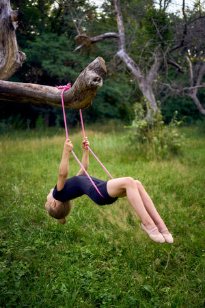 little 5-year-old gymnast in a black minimalist jumpsuit swings on a gymnastic rope on a tree