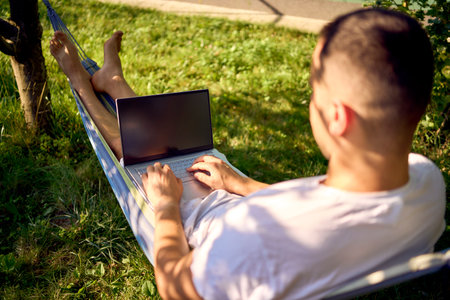a man works on a laptop in a hammock in the garden, next to his pet dogの写真素材