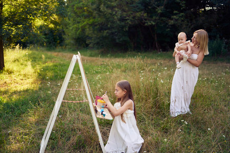mother and baby sister watch as a pre-teen girl in a summer dress paints a picture in the garden on an easelの写真素材