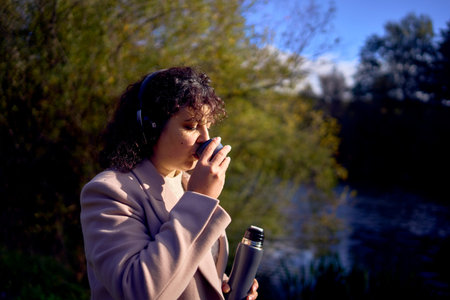 a middle-aged woman in wireless headphones drinking coffee, tea from a flask on a walk in the autumn...