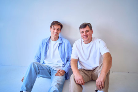 portrait of a middle-aged man  and his adult son sitting by the wall on a white background
