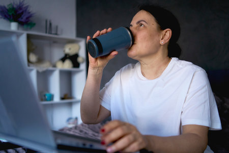 Person works from home on a laptop and drinks coffee from a thermos mugの写真素材