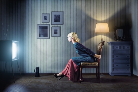 Young  woman sitting on a chair in vintage interior  and watching retro tv  She is very astonished while watching tv in dark room の写真素材